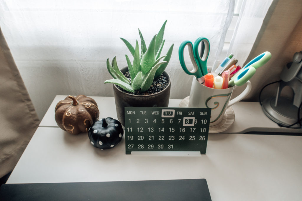 Desk with aloe plant, green magnetic desk calendar, pencil holder, brown pumpkin with gold swirls, and black pumpkin with silver polka dots